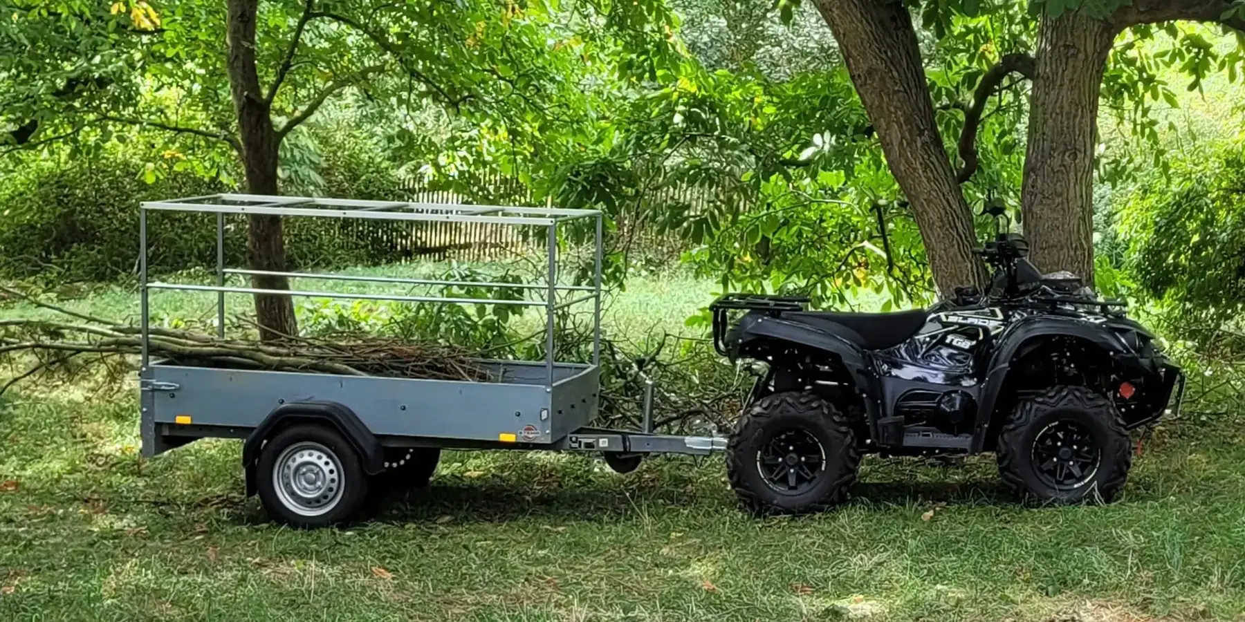 Quad mit beladenem Gitteranhänger nach Baumfällung und Ausästen auf einem Grundstück