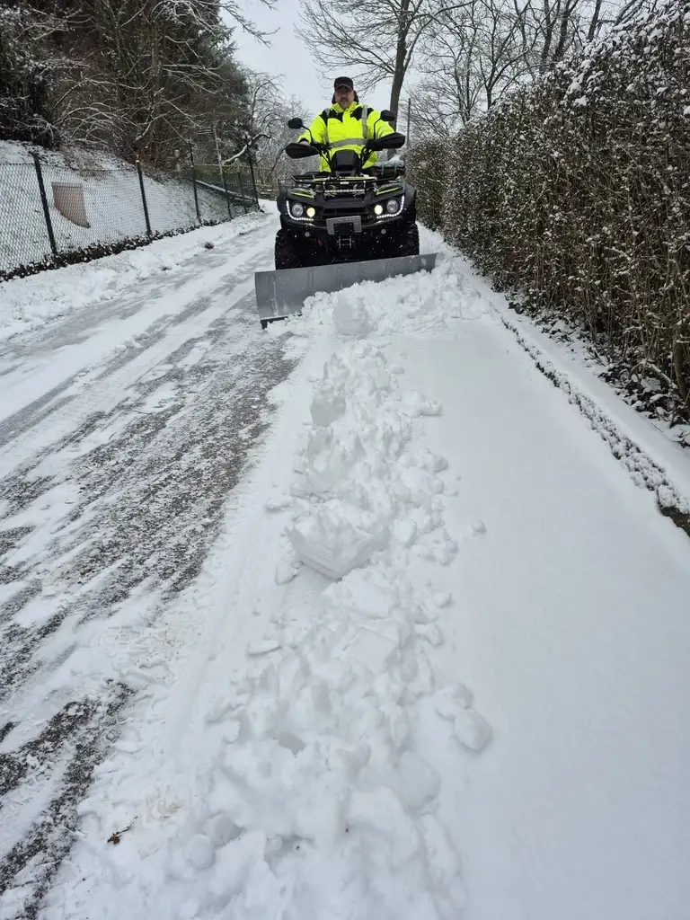 Mitarbeiter in Warnjacke räumt mit Quad und Schneeschild einen verschneiten Weg